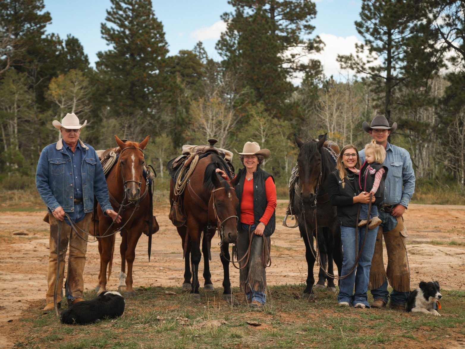 Family of cowboys and cowgirls standing with their horses in a rustic outdoor setting, featuring two adults and two children, along with a dog lying on the ground. The group is dressed in traditional Western attire, showcasing a connection to ranch life and equestrian culture.