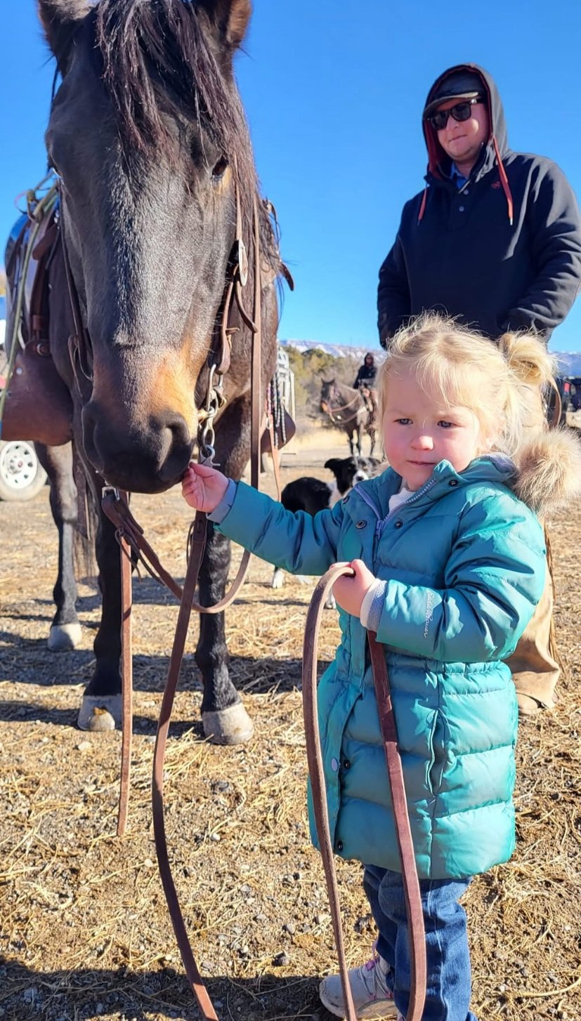 Tayler+VanWinkle++VanWinkle+Ranch+Colorado+-+853BEB08-7243-45E3-99AF-75C324F60649 A young girl in a teal coat gently holds the reins of a brown horse, while a man in a black hoodie and sunglasses stands in the background. The scene is set outdoors with clear blue skies, showcasing a rural environment.