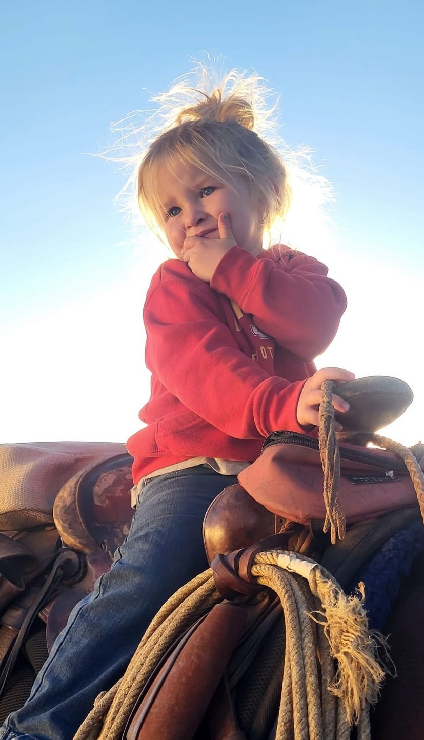 Tayler+VanWinkle++VanWinkle+Ranch+Colorado+-+9B99EFA4-8136-4F87-B2F1-B2277AA184AF Young child sitting on a saddle with a thoughtful expression, wearing a red sweatshirt and blue jeans, against a bright sky backdrop. The image captures a playful moment in a rural setting.